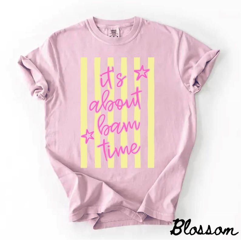 May include: A pale pink t-shirt featuring yellow vertical stripes and the pink text "It's about bam time". Two pink stars are positioned near the text. The word "Blossom" is in black script at the bottom right.