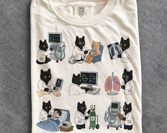 Op de afbeelding: Een ivoorkleurig t-shirt met een grafische print van zwarte katten verkleed als ademhalingstherapeuten die diverse medische taken uitvoeren, zoals het bedienen van beademingsapparatuur, monitoren controleren en röntgenfoto's bekijken.