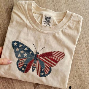 以下が含まれることがあります： アイボリー色のComfort Colors Tシャツ。愛国的な蝶のデザインが特徴です。蝶の羽は赤と白のストライプと、白い星が付いた青い部分で構成されており、アメリカ国旗を連想させます。クルーネックと半袖です。
