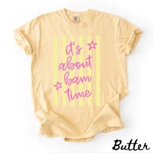 May include: Pale yellow t-shirt featuring the text "It's about bam time" in pink script with star accents, set against vertical yellow stripes. The word "Butter" is printed in a cursive font at the bottom right corner of the shirt.