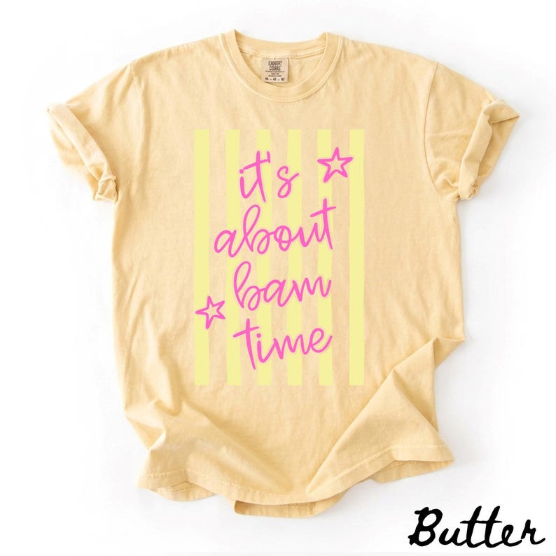 May include: Pale yellow t-shirt featuring the text "It's about bam time" in pink script with star accents, set against vertical yellow stripes. The word "Butter" is printed in a cursive font at the bottom right corner of the shirt.