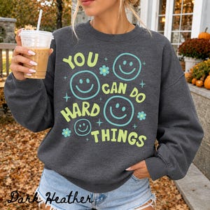 May include: A dark heather gray crewneck sweatshirt featuring the text YOU CAN DO HARD THINGS in light green and light blue. The design includes four smiling faces and small flower and star icons printed in light blue and light green ink.
