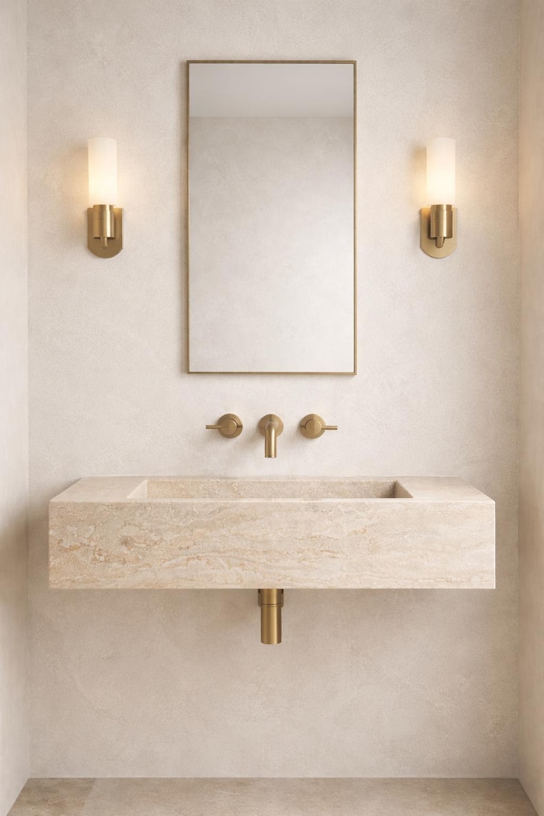 Op de afbeelding: Een moderne badkamerset met een rechthoekige stenen wastafel, messing armaturen en een rechthoekige spiegel. Twee wandlampen flankeren de spiegel en zorgen voor zachte verlichting. Het ontwerp is minimalistisch en elegant, met een neutraal kleurenpalet.