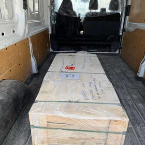 Op de afbeelding: Een grote, rechthoekige houten kist verpakt in doorzichtig plastic, vastgezet met groene banden en beschreven met handgeschreven tekst. De kist staat in een witte bestelwagen.