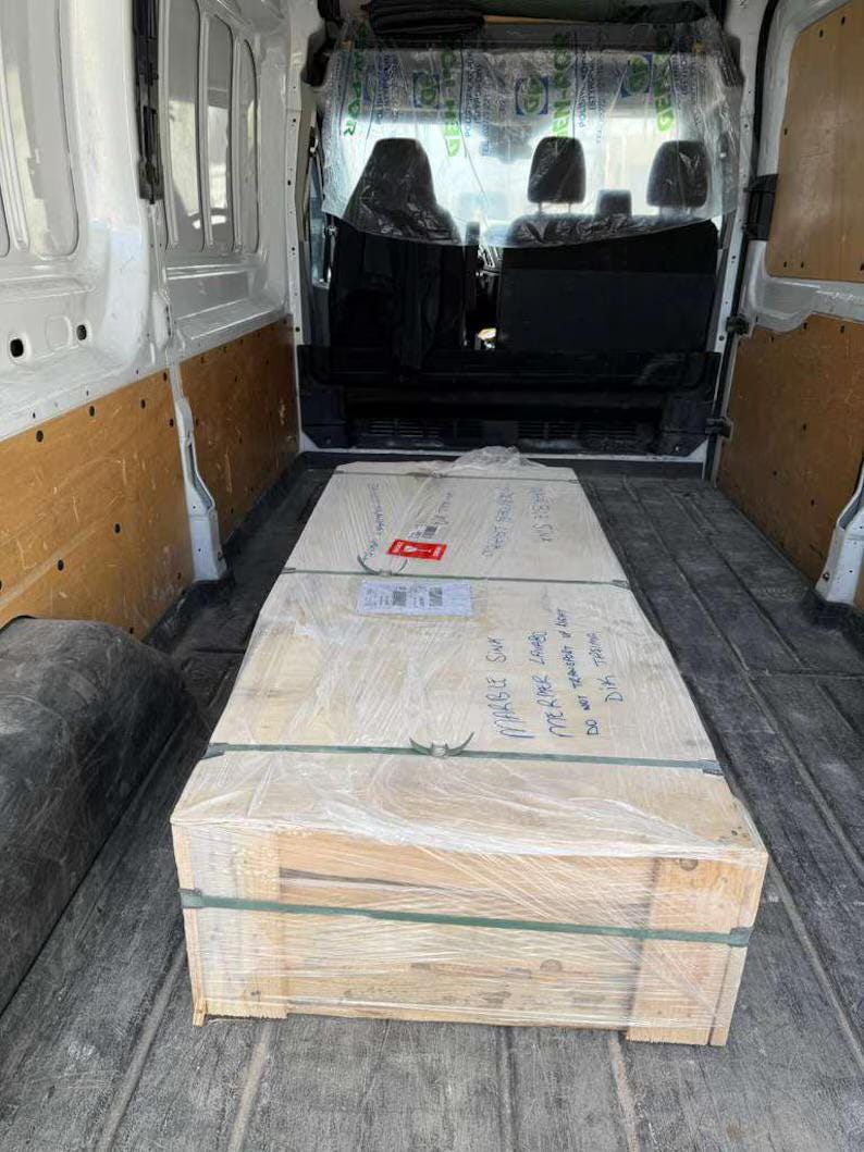 Op de afbeelding: Een grote, rechthoekige houten kist verpakt in doorzichtig plastic, vastgezet met groene banden en beschreven met handgeschreven tekst. De kist staat in een witte bestelwagen.
