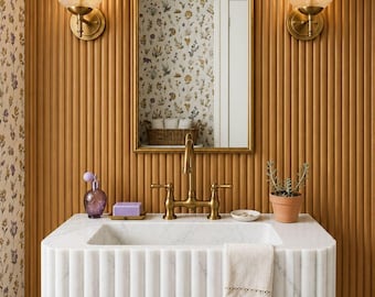 May include: A white marble wall-mounted sink with a fluted front, featuring a brass tap, a purple soap bar, a purple perfume bottle, a small potted plant, and a beige towel. Above, an arched mirror is flanked by two ribbed glass wall sconces.