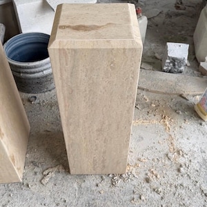 May include: A tall, rectangular pedestal made of light beige stone with a smooth, polished finish. The top surface is flat with beveled edges, and the sides feature natural, subtle veining patterns throughout the stone structure.
