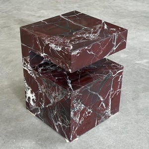 May include: A cubic side table crafted from deep red marble with prominent white veining. The structure features a solid base and a cantilevered top section, creating a distinct horizontal gap between the two main blocks of the stone furniture piece.