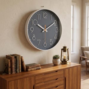 May include: A round, gray wall clock with white numerals and hands, and an orange second hand. The clock has a white frame and is mounted on a beige wall. The word "QUARTZ" is printed on the clock face. The clock is part of a home decor theme.