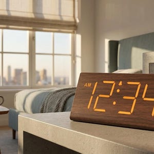 May include: A wooden digital alarm clock displaying 12:34 AM. The clock has a brown finish and a rectangular shape with a slanted top. The numbers are illuminated in a warm orange color. The clock is on a bedside table.