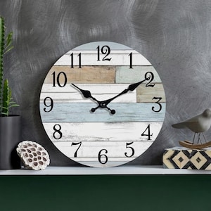 May include: Round wall clock with a distressed wood-look face in shades of blue, white, and tan. Black hands and numerals. The clock is displayed on a white shelf with decorative items, including a small bird figurine.