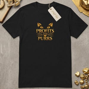 Może przedstawiać: Czarny t-shirt z krótkim rękawem ze złotą grafiką przedstawiającą kocie oczy, wąsy i małe serce. Poniżej oczu znajduje się złoty napis "PROFITS AND PURRS". Do koszulki przymocowana jest biała metka z napisem "P&P Est.".