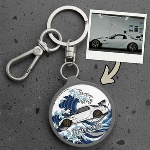 Op de afbeelding: Een ronde sleutelhanger met een zilverkleurige metalen ring en karabijnhaak. De hanger bevat een grafische illustratie van een witte sportwagen boven een blauw gestileerd golfpatroon met de tekst Drive safe in een wit cursief lettertype.