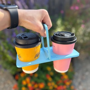 Puede incluir: Un portavasos de plástico azul claro con un asa central que sostiene dos vasos desechables. Un vaso es amarillo con tapa negra y el otro es rosa con tapa negra. El soporte presenta un pequeño logotipo en relieve en la superficie lateral.
