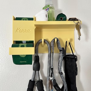 May include: A pale yellow wall-mounted organiser featuring a shelf, a side compartment for waste bag rolls, and four hooks. The side is embossed with the name Konna. The shelf holds a spray bottle, white case, and keys, while the hooks hold leashes.