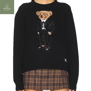May include: Black knit sweater featuring a bear wearing a suit. The bear is in shades of brown, black, and white. The sweater has the letters "RL" embroidered on the lower right side. The sweater is paired with a brown plaid skirt.
