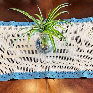 May include: A handmade, rectangular doily with a white center and a blue border. The doily has a geometric pattern and is topped with a glass vase holding a green plant. The vase is filled with blue decorative stones.