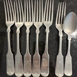 May include: A collection of antique silver forks and a spoon. The forks have four tines and ornate handles with engraved initials. The spoon has a rounded bowl and a similar handle design. The silverware is arranged on a dark surface.