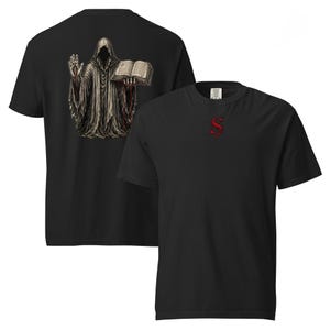 May include: Black t-shirt featuring a detailed graphic of a robed figure holding a book on the back. The front of the shirt displays a red letter "S". The design is intricate, with a gothic or fantasy theme.
