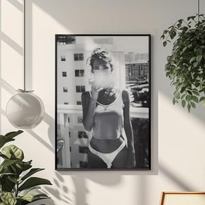 Puede incluir: Fotografía enmarcada en blanco y negro de una persona en un balcón, exhalando humo. La persona lleva una camiseta sin mangas y ropa interior. El fondo muestra un horizonte urbano. La habitación tiene plantas y una silla de madera.