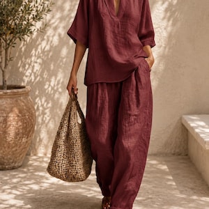 May include: A deep burgundy two-piece set consisting of a V-neck top with elbow-length sleeves and matching loose-fitting trousers with a cuffed hem. The outfit is paired with a woven tan tote bag and brown leather strappy sandals.