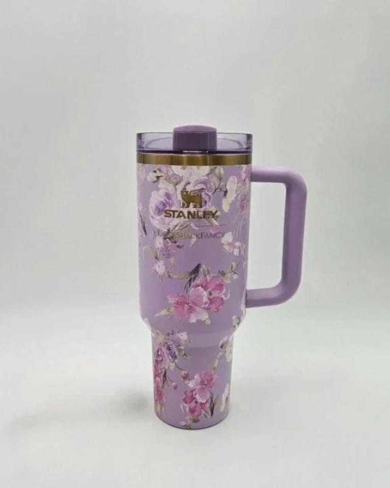 Puede incluir: Un vaso Stanley lavanda con un dise&ntilde;o floral de flores rosas y blancas. El vaso tiene un asa, una tapa y el logotipo de Stanley. Tambi&eacute;n se ve el texto "SHACKFANCY".