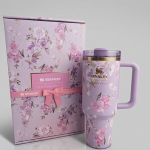Puede incluir: Un vaso Stanley lavanda con asa y dise&ntilde;o floral, junto a una caja de regalo a juego. El vaso y la caja presentan flores rosas y moradas. La caja de regalo tiene una cinta rosa y una tarjeta que dice "Stanley Loveshackfancy".