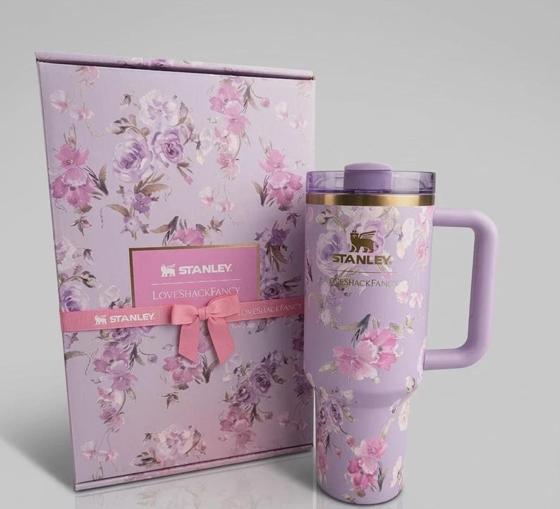 Puede incluir: Un vaso Stanley lavanda con asa y dise&ntilde;o floral, junto a una caja de regalo a juego. El vaso y la caja presentan flores rosas y moradas. La caja de regalo tiene una cinta rosa y una tarjeta que dice "Stanley Loveshackfancy".