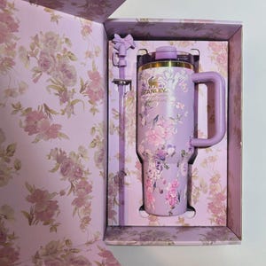 Puede incluir: Un vaso Stanley lavanda con un dise&ntilde;o floral, una pajita a juego y un asa, presentado en una caja con estampado floral. El vaso presenta el logotipo de Stanley y un borde dorado. La caja y la pajita tienen un detalle en forma de lazo.