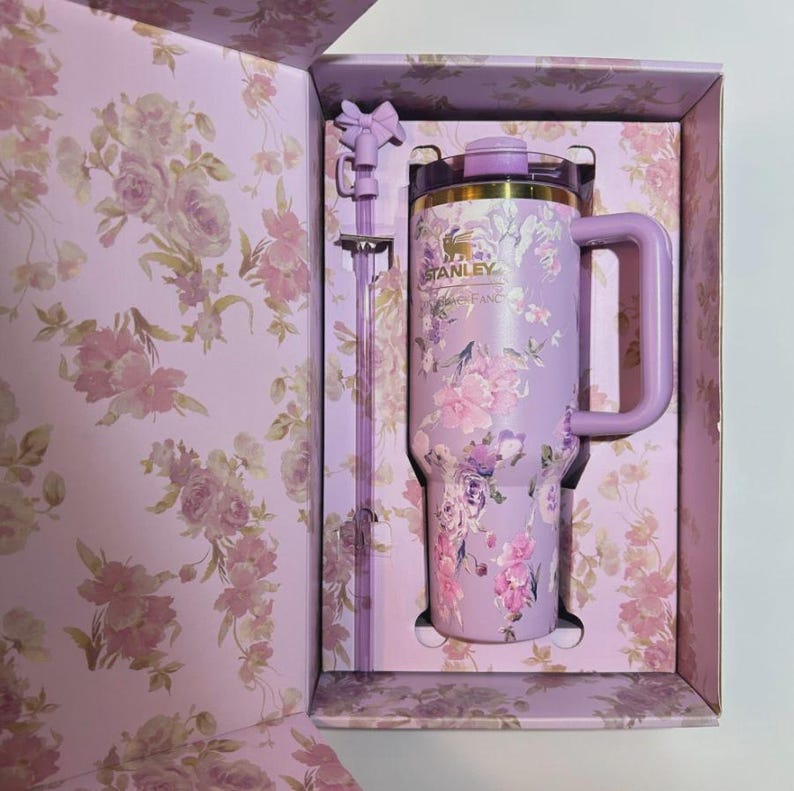 Puede incluir: Un vaso Stanley lavanda con un dise&ntilde;o floral, una pajita a juego y un asa, presentado en una caja con estampado floral. El vaso presenta el logotipo de Stanley y un borde dorado. La caja y la pajita tienen un detalle en forma de lazo.