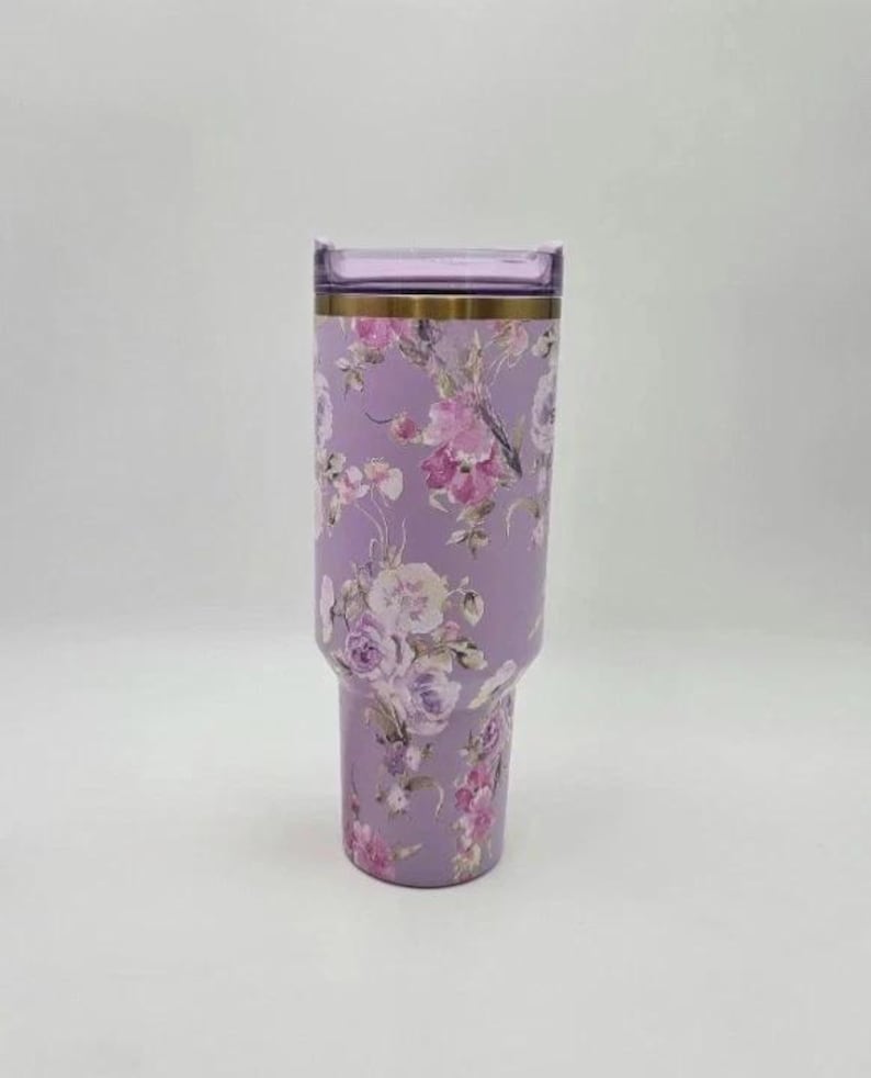 Puede incluir: Un vaso de color lavanda con un dise&ntilde;o floral. El vaso presenta una banda dorada cerca de la parte superior y una tapa transparente. El patr&oacute;n floral incluye flores rosas, moradas y blancas. El vaso est&aacute; dise&ntilde;ado para contener unos 887 ml.