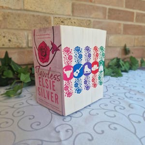 Könnte beinhalten: Ein Stapel Bücher mit dem Titel "Flawless" von Elsie Silver. Das Buchcover zeigt eine rosa Rose und der Buchrücken farbenfrohe Illustrationen eines Stiers, einer Gitarre, eines Cowboyhuts und anderer Symbole. Die Bücher liegen auf einer gemusterten Oberfläche.