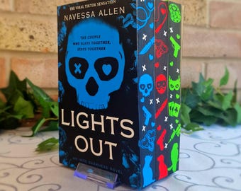 May include: A book titled "LIGHTS OUT" by Navessa Allen, with a blue skull design on the cover. The spine features colorful illustrations. The text reads "THE VIRAL TIKTOK SENSATION" and "THE COUPLE WHO SLAYS TOGETHER, STAYS TOGETHER."