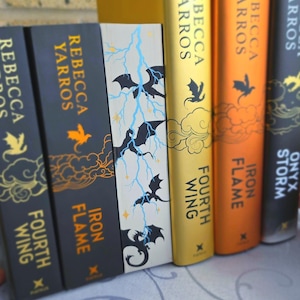 May include: A collection of hardback books by Rebecca Yarros, including Fourth Wing, Iron Flame, and Onyx Storm. The spines feature gold and orange text, dragon silhouettes, and cloud motifs. One book edge displays a hand-painted blue lightning design.