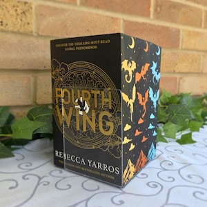 May include: A black book titled Fourth Wing by Rebecca Yarros with gold foil lettering and circular graphic details. The side panel features colourful dragon silhouettes in yellow, orange, and blue against a black background with clouds and mountains.