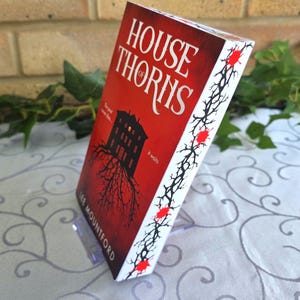 May include: A paperback book titled House of Thorns by Lee Mountford with a red cover featuring a black silhouette of a house with roots, the text The past never dies... it waits, and a white spine decorated with black thorny vines and red spots.