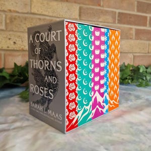 May include: A decorative box set of 'A Court of Thorns and Roses' books. The box has a grey spine with the title and author's name, and patterned sides with roses, moons, mountains, and flames.