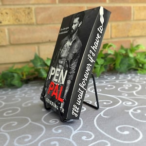 May include: A rectangular black book block titled Pen Pal by J.T. Geissinger, featuring a black and white portrait of a man on the cover. The side spine displays the text I'll wait forever if I have to with a white fountain pen nib icon.