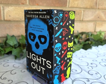May include: A paperback book titled Lights Out by Navessa Allen with a black cover featuring a blue skull graphic and the text The couple who slays together, stays together. The book spine displays a pattern of colorful icons including skulls, knives, and hearts against a black background.