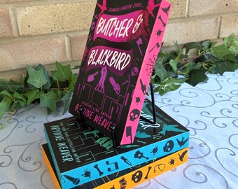 May include: A stack of three books titled Butcher & Blackbird by Brynne Weaver. The top book is pink and black with a silhouette of two people. The middle book is teal with black icons, and the bottom book is orange with black icons of bones and tools.