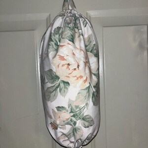 Puede incluir: Una bolsa de tela con un estampado floral de grandes rosas melocotón y blancas y hojas verdes. La bolsa cuelga de un gancho blanco. La bolsa está fruncida en la parte superior con un cordón y tiene una pequeña abertura en la parte inferior.