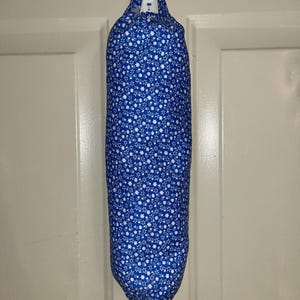 May include: A blue and white fabric bag dispenser hanging on a white hook. The bag is cylindrical and patterned with small white floral shapes. The dispenser is hanging on a white door.