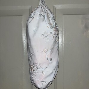 Puede incluir: Una bolsa de tela con estampado floral en tonos blancos, rosas y grises, colgada de un gancho blanco. La bolsa tiene un cierre de cordón en la parte superior y una pieza de plástico en la parte inferior. Para guardar.