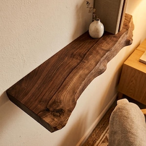 May include: A dark brown, live edge wooden shelf mounted on a white wall. The shelf holds two books, a small white vase, and a sprig of greenery. The shelf has a natural, organic shape and texture.