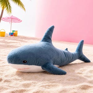 May include: A plush shark toy with a dark blue top and a white underside. The stuffed animal features a prominent dorsal fin, side fins, a tail, and embroidered eyes. The shark has a wide mouth with visible white teeth and small gill slits.