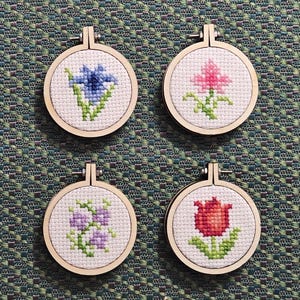 May include: Four small circular wooden embroidery hoops containing white Aida fabric with cross-stitched floral designs. The patterns feature a blue flower, a pink flower, a cluster of purple blossoms, and a red tulip, each with green stems and leaves.