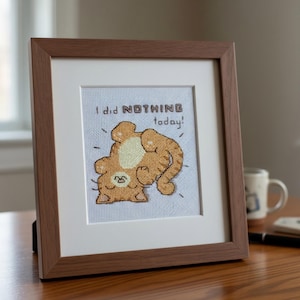 Op de afbeelding: Een ingelijst kruissteek borduurwerk met een oranje cyperse kat die op de rug ligt, met de tekst "I did NOTHING today!" geborduurd in bruine draad. Het kunstwerk zit in een eenvoudige bruine houten lijst met een witte passe-partout.
