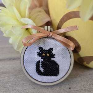 May include: A small circular wooden embroidery hoop containing white fabric with a cross-stitched black cat featuring yellow eyes. A light peach-coloured satin ribbon is tied in a bow at the top of the wooden frame, resting above the cat design.