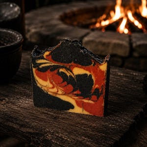 May include: A rectangular bar of soap featuring a marbled swirl pattern in black, deep red, and warm orange. The soap has a textured, uneven top edge and a smooth front face, showcasing a distinct blend of dark and vibrant colours throughout the bar.