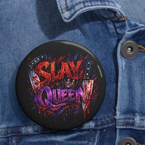 May include: A round black button with the words "SLAY QUEEN" in red and purple, with a crown and two knives. The button has a glossy finish and is set against a denim jacket.